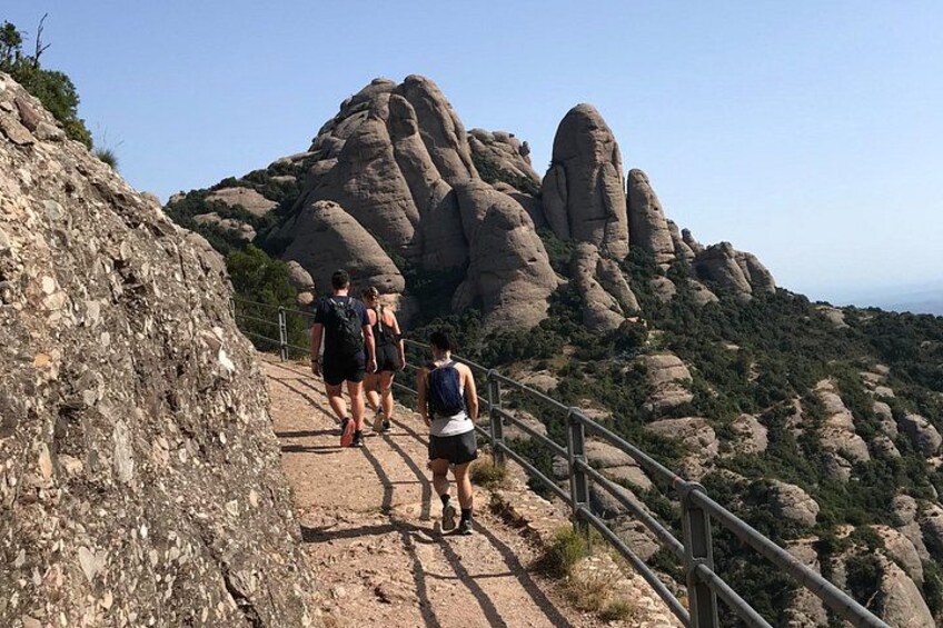 A group of hikers on a rocky trail with a metal railing on the right, surrounded by large rock formations and sparse vegetation. 4
