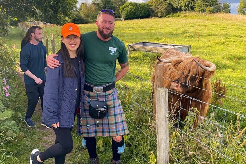 A person in a kilt and cap standing next to a cow in a fenced field. 2