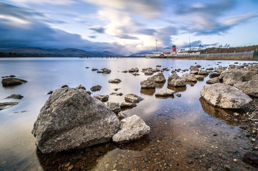 A calm lake with large rocks in the foreground and a boat with a red and white cabin in the distance. 4