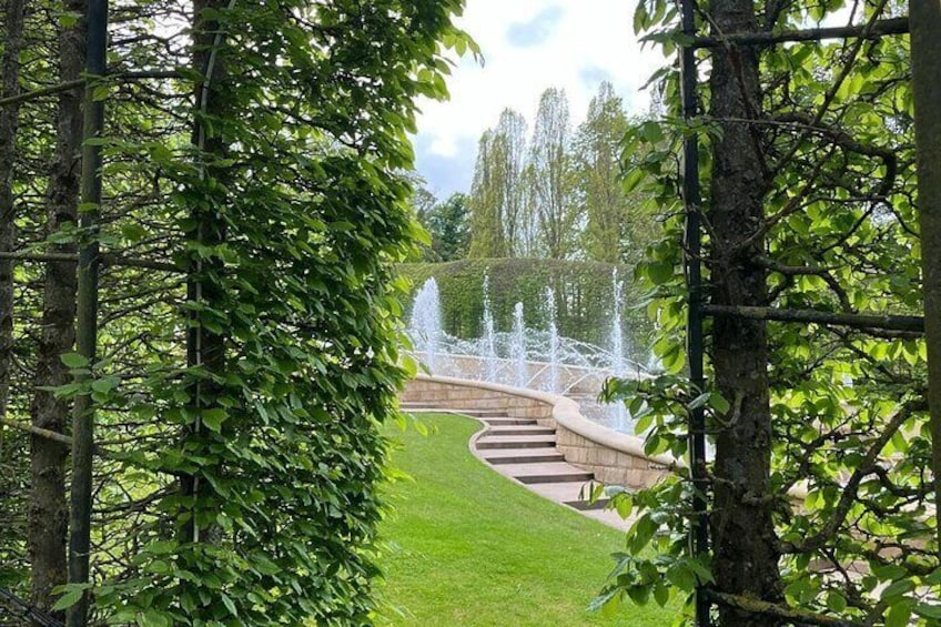 A curved stone pathway with a fountain in the background, surrounded by lush green trees. 3