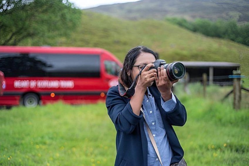 A person taking a photograph with a camera, a red bus in the background, and a grassy field. 3