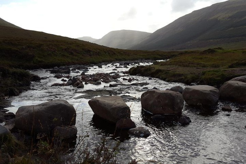 Un río con rocas grandes, colinas cubiertas de hierba y montañas brumosas al fondo. 1