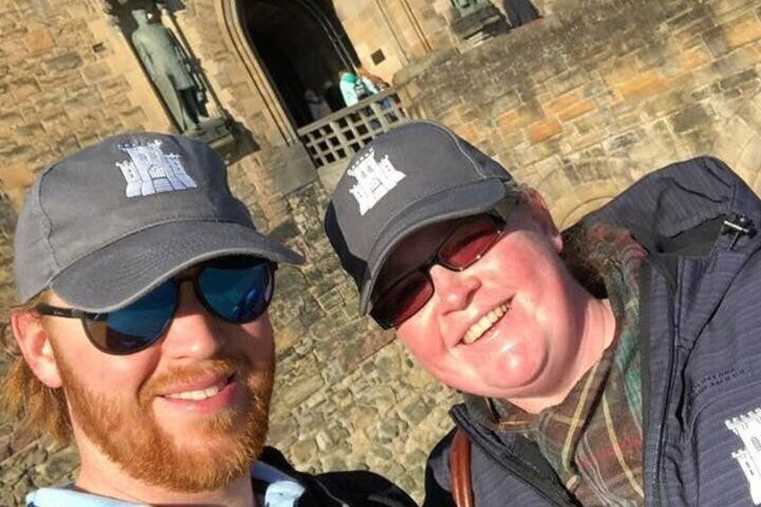 Two people wearing matching caps with a logo, standing in front of a stone building with a door and a window. 2
