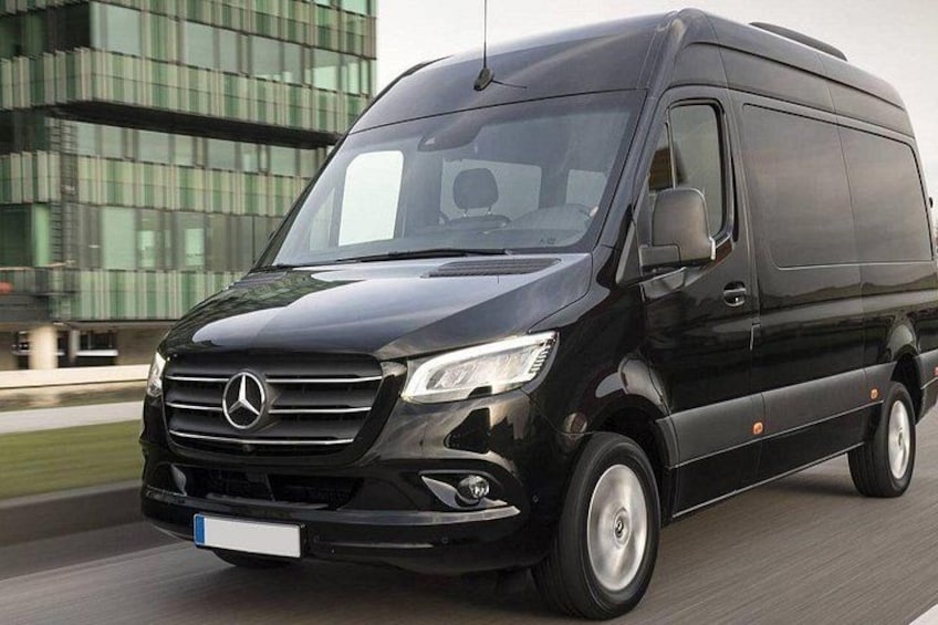 Un fourgon Mercedes noir roule sur une route, avec un bâtiment moderne en arrière-plan. 2