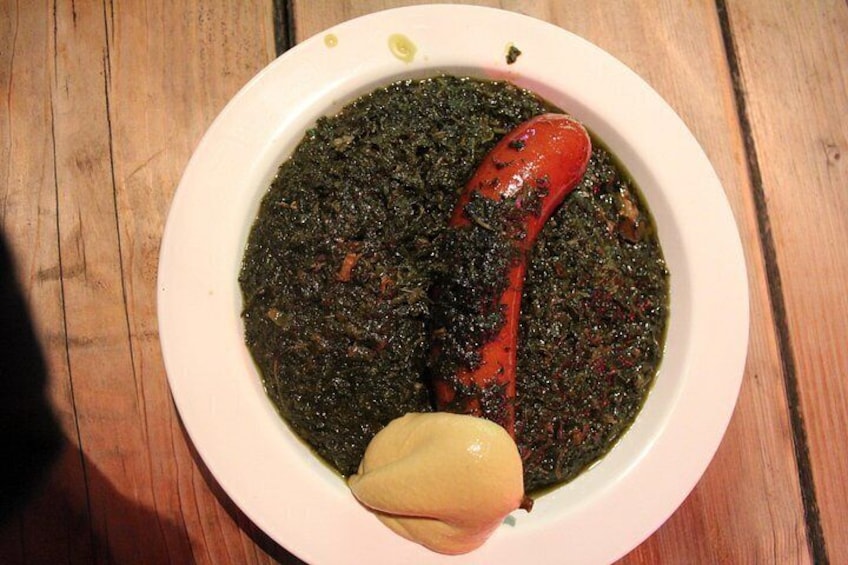 A bowl of dark green stew with a red sausage and a piece of garlic. 4
