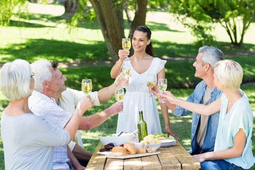 A group of people toasting with wine glasses around a picnic table in a park. 1