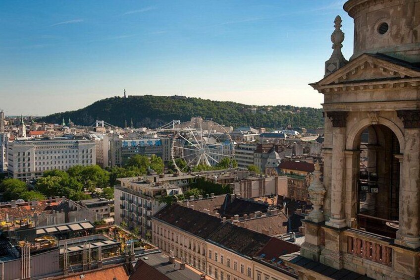 Une vue urbaine avec une grande roue bien visible, des bâtiments d’architecture historique et une colline en arrière-plan. 2