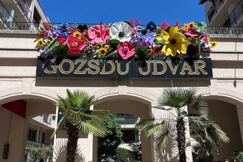 A building entrance with a sign reading 'GOZSDU UDVAR' and a floral arrangement above it. 5