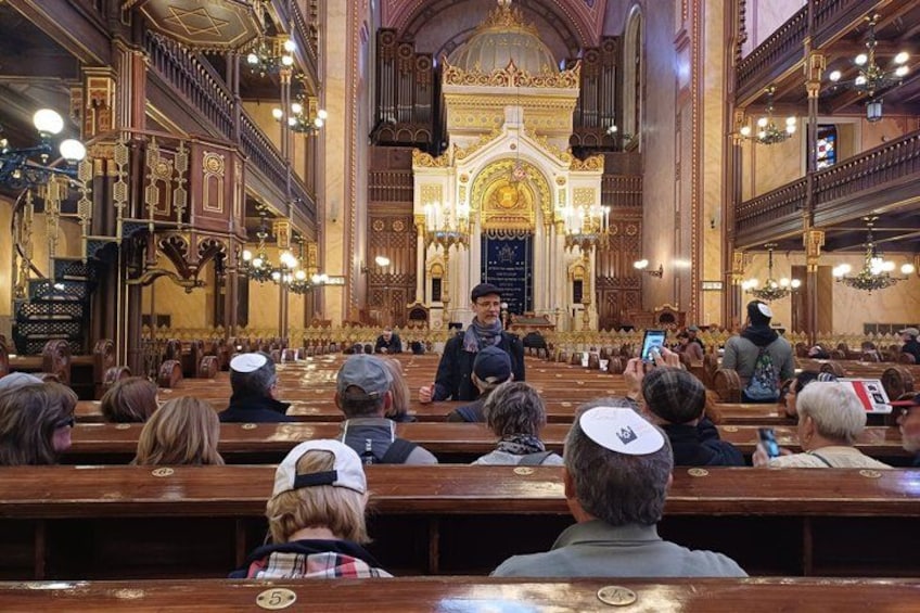 A synagogue interior with ornate woodwork, a central podium, and attendees seated on wooden benches. 1