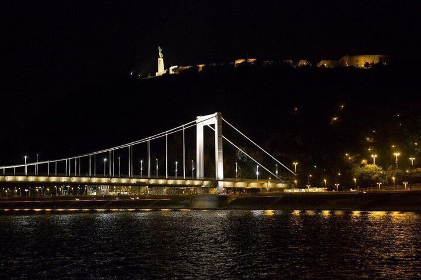 A nighttime view of a bridge with lights, a body of water, and a distant illuminated structure on a hill. 5
