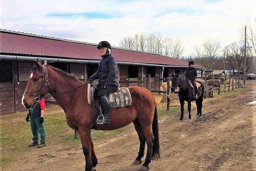 A person riding a horse on a dirt path, with another horse and rider in the background. 1