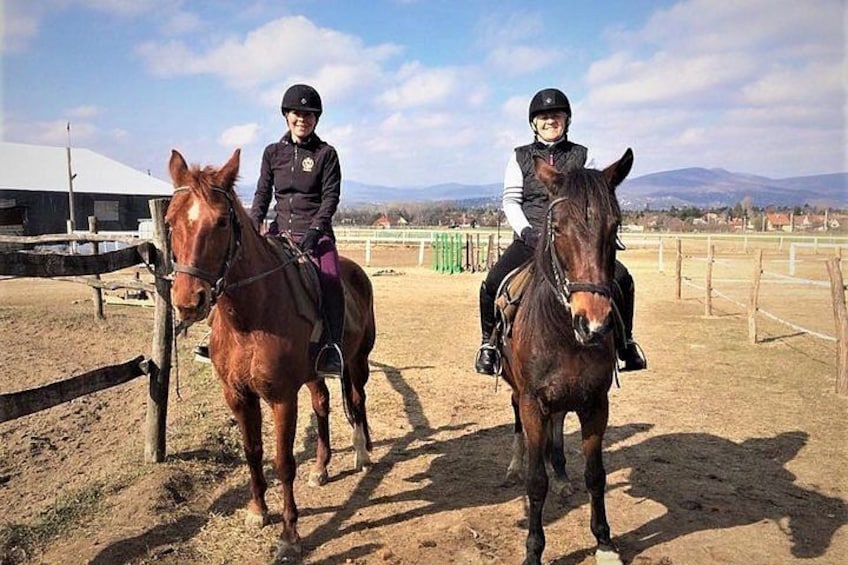Two people riding horses in an outdoor arena with a fence, a building, and a clear sky. 2