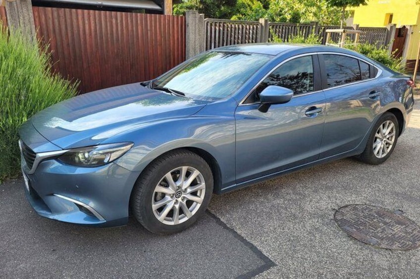 A blue sedan parked on a driveway with a wooden fence and greenery in the background. 2