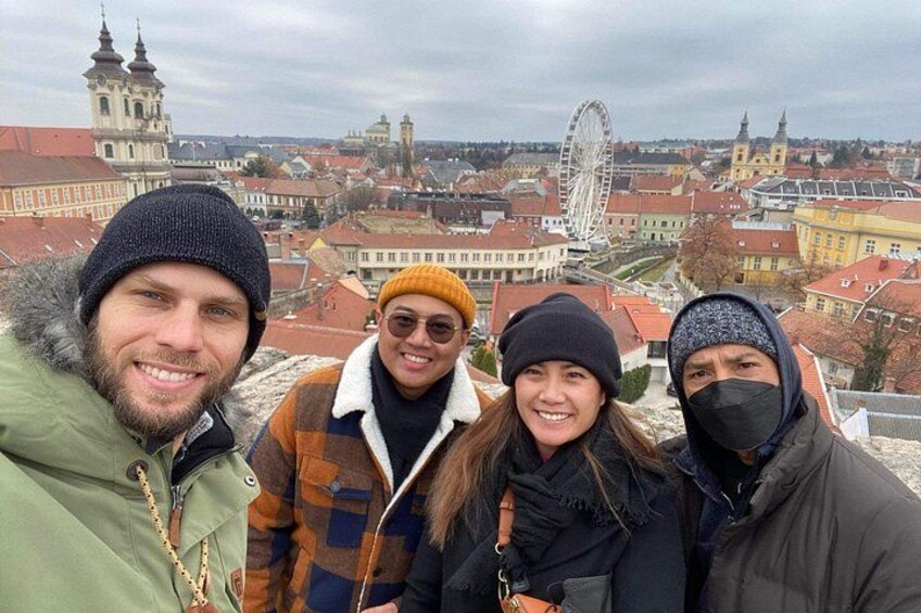 A group of people standing on a high vantage point overlooking a cityscape with historic buildings and a large Ferris wheel. 2