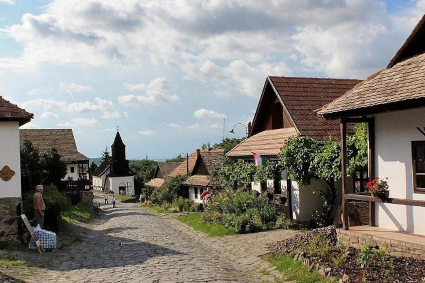 A cobblestone street in a village with traditional houses, a church steeple, and a person sitting on a bench. 1