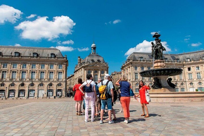 Een groep mensen staat op een plein met een fontein en historische gebouwen op de achtergrond. 3