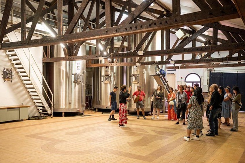 A group of people in a spacious industrial setting with metal beams, large stainless steel tanks, and a wooden floor. 5