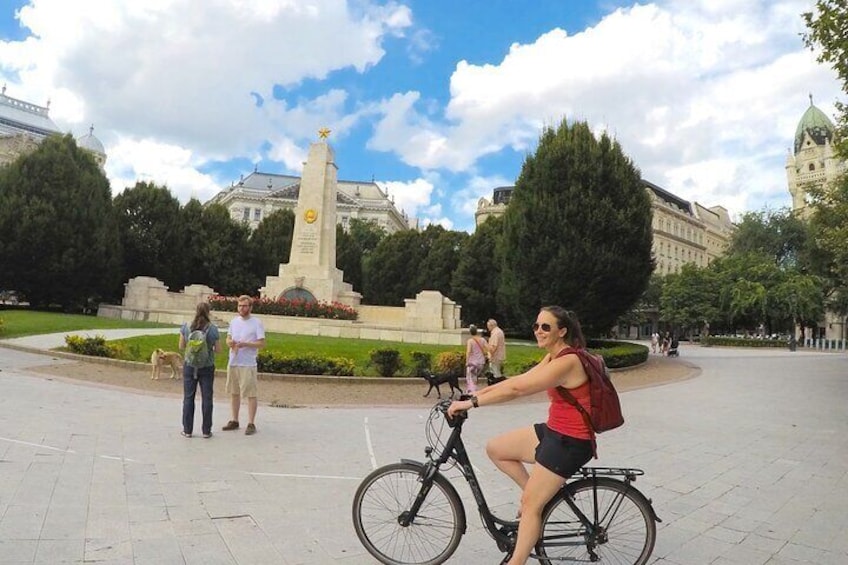 Una persona montando una bicicleta en una plaza pública con un monumento al fondo. 5