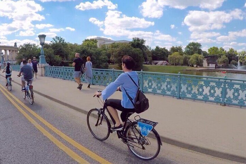 A person riding a bicycle on a bridge with a blue basket, pedestrians walking, and a fountain in the background. 3