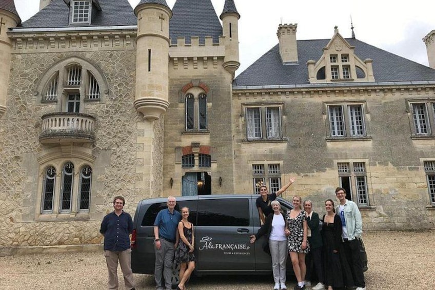 Un groupe de personnes se tient devant un grand bâtiment en pierre, doté de nombreuses fenêtres, et un véhicule est visible à proximité. 2