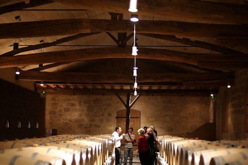 A wine cellar with wooden beams and stone walls, rows of wine barrels, and people standing in the center. 2