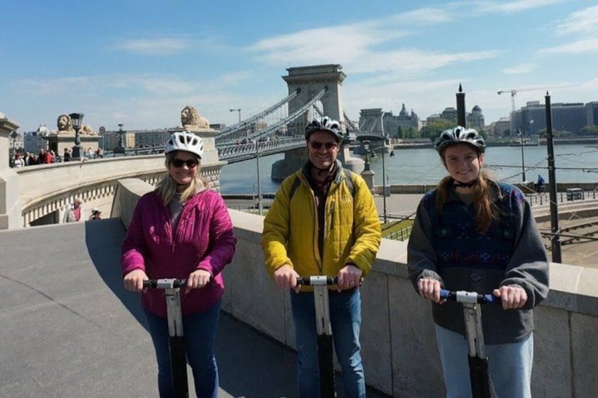 Three people on scooters near a bridge with a river and buildings in the background. 2