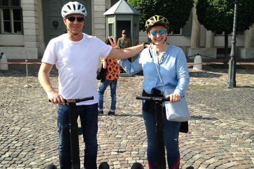 Twee personen op scooters in een geplaveid gebied, met een gebouw op de achtergrond. 3