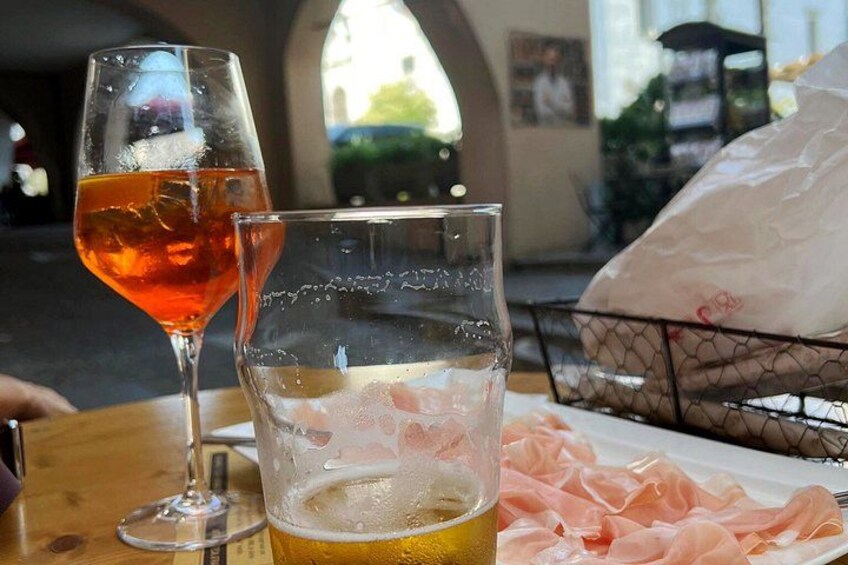 A glass of beer and a glass of rosé wine on a table with a plate of prosciutto. 4