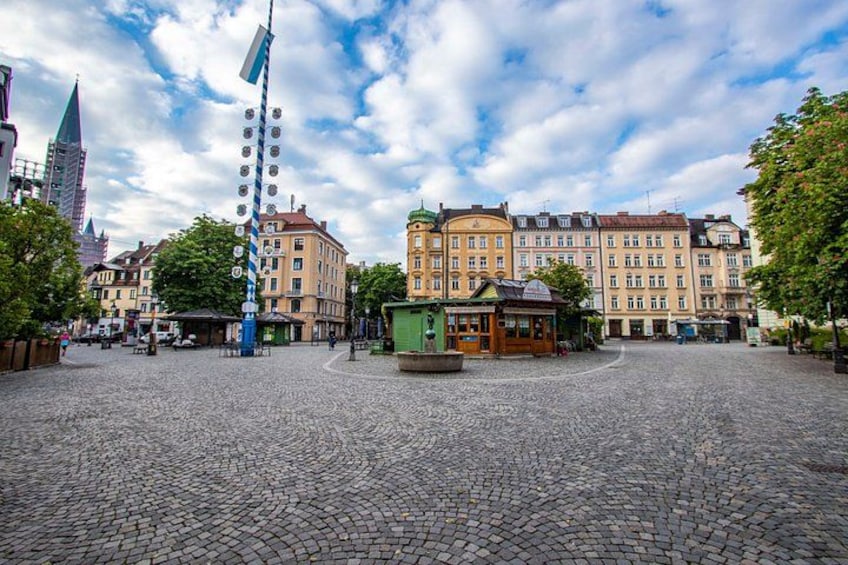 A cobblestone square with historic buildings, a tall flagpole, and a small kiosk. 1