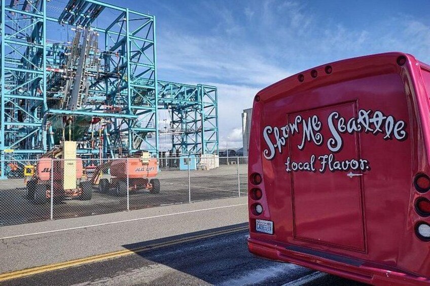 En röd buss med texten ”Visa mig Seattle” och ”lokal prägel” på baksidan, parkerad på en väg med en blå industriell byggnad i bakgrunden. 2