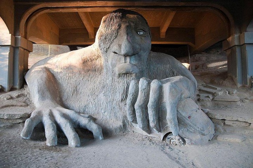 En stor stenskulptur som föreställer en liggande figur med utsträckta armar, placerad utomhus med en träkonstruktion i bakgrunden. 2