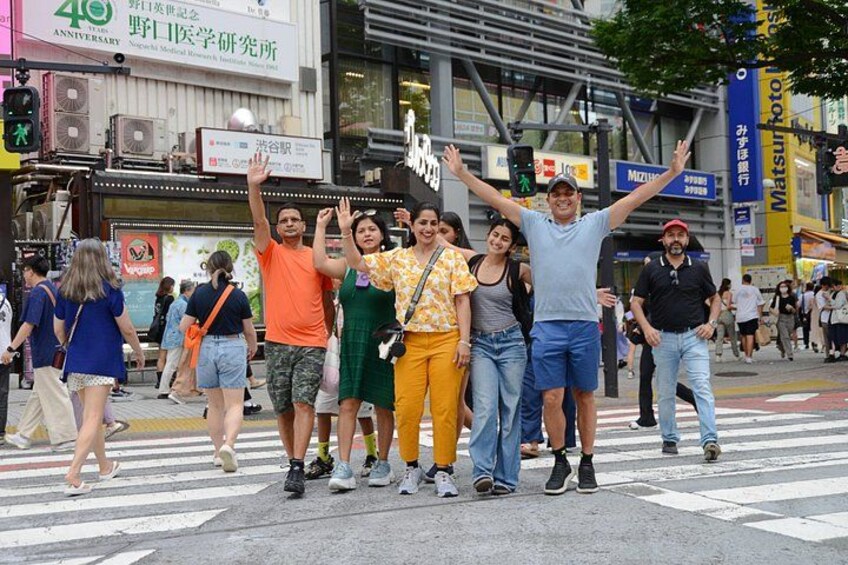 A group of people crossing a street with a crosswalk, buildings, and a signboard in the background. 1
