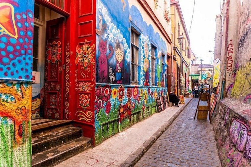 A narrow alley with colorful murals on buildings, steps leading up to a door, and a cobblestone path. 1