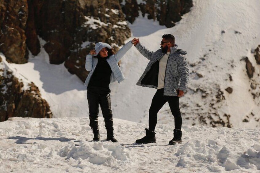Two people standing on a snowy mountain with rocky cliffs in the background. 4