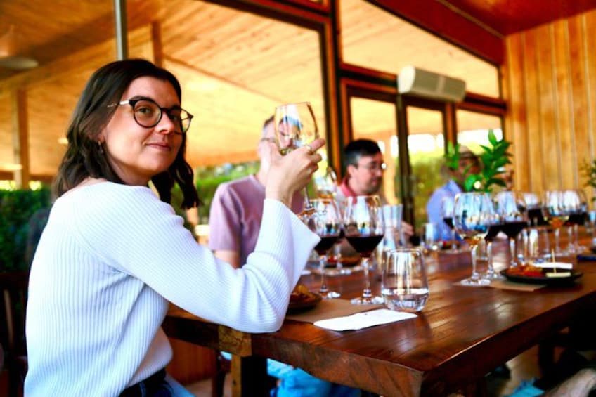 A person holding a wine glass at a wooden table with several other people seated, suggesting a social gathering or event. 4