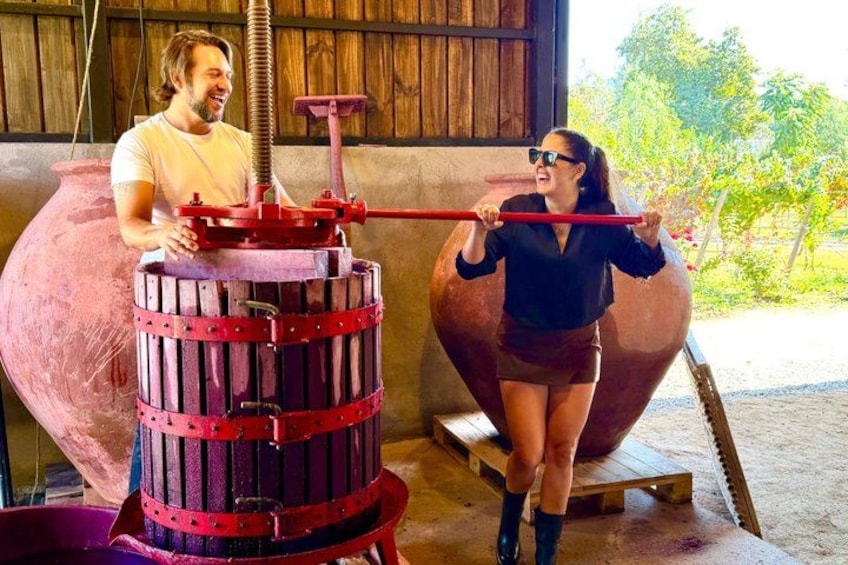A person operating a large red mechanical press in a rustic setting with wooden structures and a large copper vessel in the background. 5