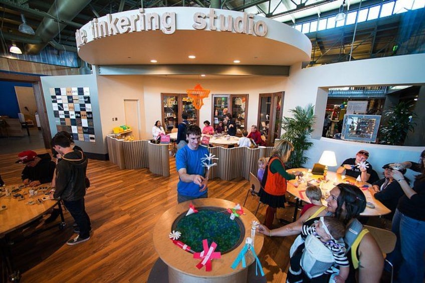 Et livligt indre rum med en buet disk med teksten 'linking studio', omgivet af borde hvor folk sidder og er engagerede i forskellige aktiviteter. 3