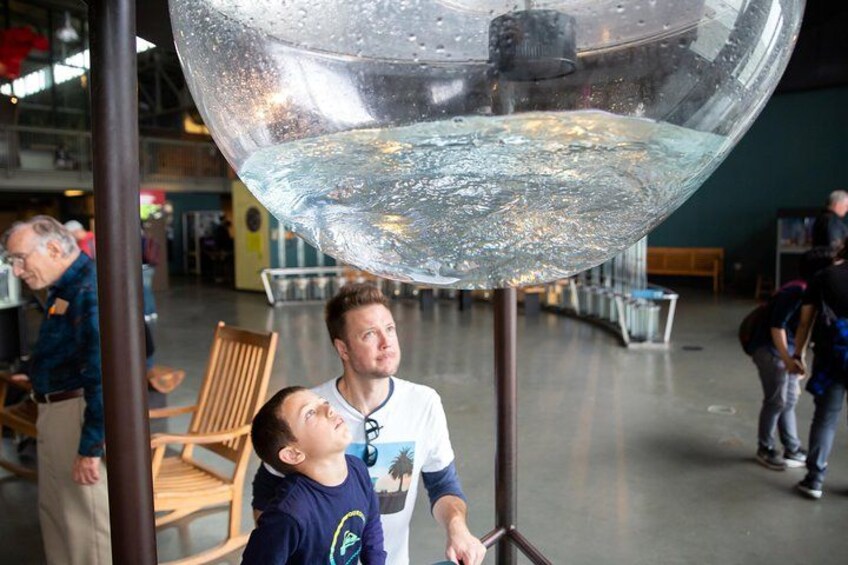 A large transparent sphere with a reflective interior, suspended from a metal pole, in a public indoor space with seating and other people in the background. 4