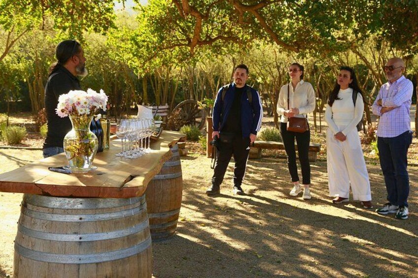 A group of people standing around a wooden table with barrels, a vase of flowers, and a bench in a wooded area. 5