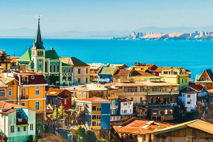 Une ville côtière aux bâtiments aux couleurs vives, une église surmontée d’un toit vert et une vue dégagée sur la mer et le lointain horizon urbain. 1