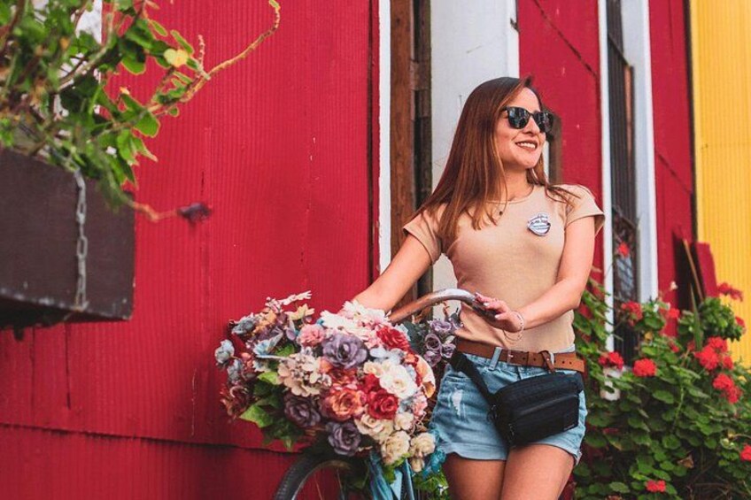 A person standing next to a bicycle with a floral basket, wearing a beige top and denim shorts, with a red building in the background. 3