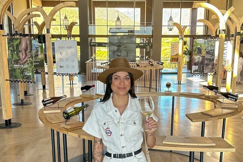 Una persona con camisa y sombrero blancos sostiene una copa de vino en un museo con mesas de madera y vitrinas. 5