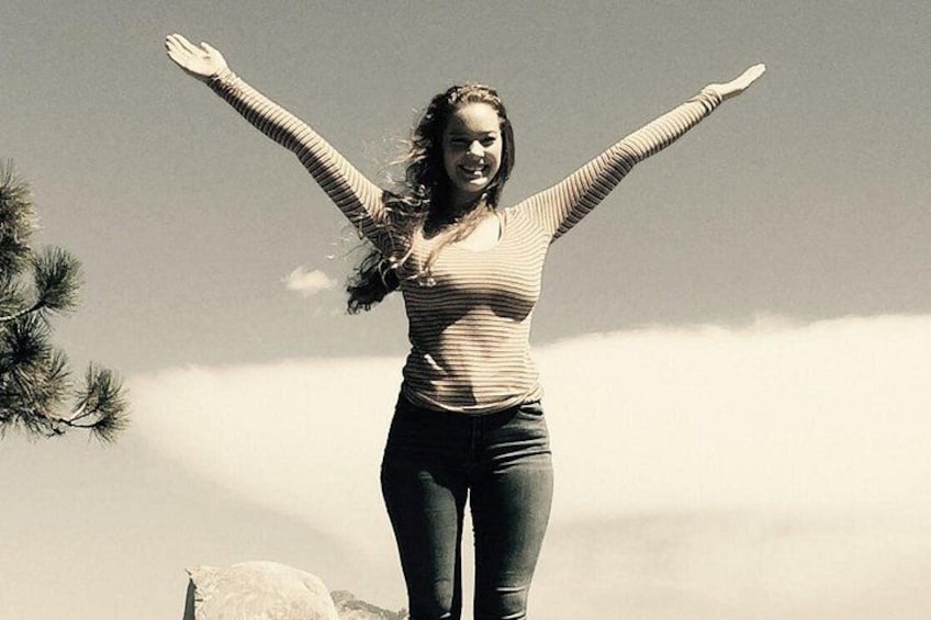 A person standing with arms raised, wearing a striped top and jeans, outdoors with a clear sky and a pine tree in the background. 3