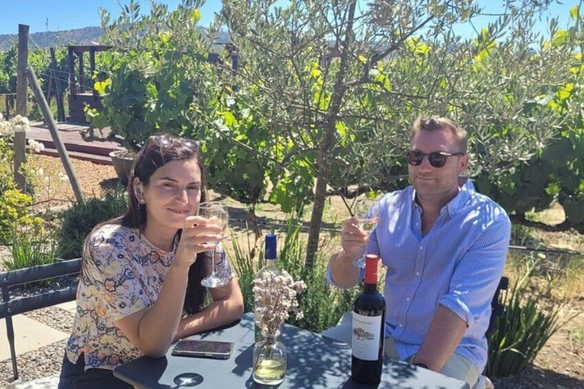 Two people sitting at a table outdoors with a bottle of wine and a glass of wine. 1