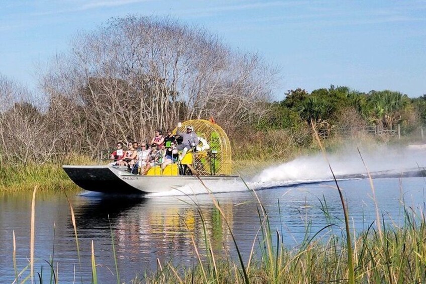 A boat with people on it, moving through water with vegetation on the banks. 1