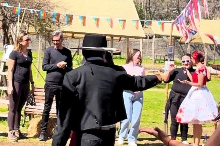 A person in a black suit and hat, possibly officiating, with a group of people in a casual outdoor setting. 2