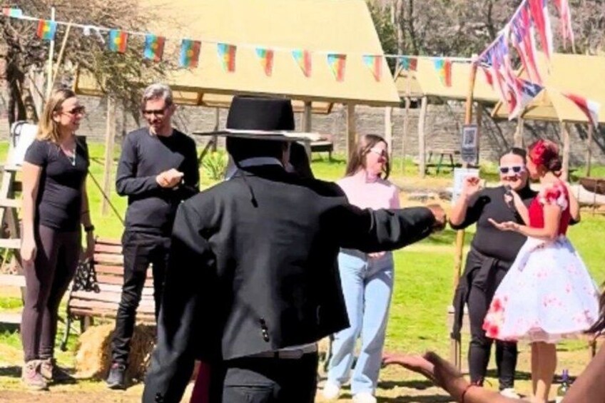 A person in a black suit and hat, a woman in a red dress, and others in a casual outdoor setting with tents and flags. 2