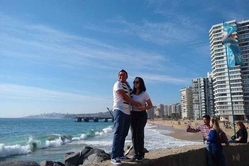A couple standing on a rocky beach with ocean waves, a pier in the background, and high-rise buildings. 2