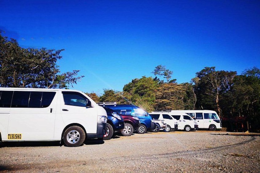 A line of vehicles parked on a gravel surface with trees in the background. 1
