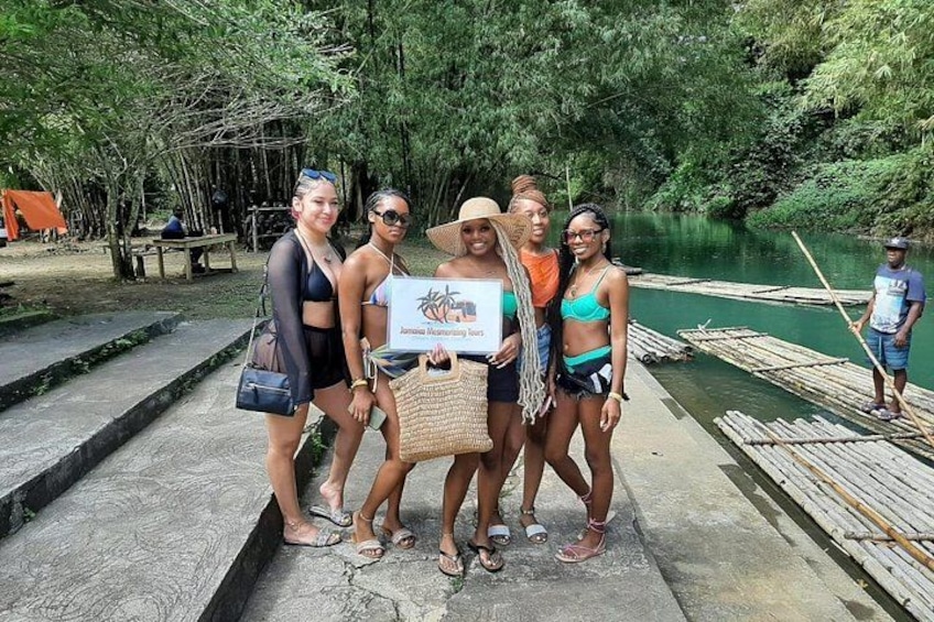 A group of people standing on a concrete path near a river with boats and a person rowing a boat. 1